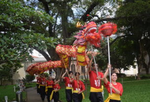 Nove pessoas vestindo camisetas vermelhas e faixas amarelas executam dança do dragão em caminho de parque com árvores e gramado. Dragão vermelho e amarelo é sustentado por varas, com uma participante segurando uma esfera vermelha na frente.