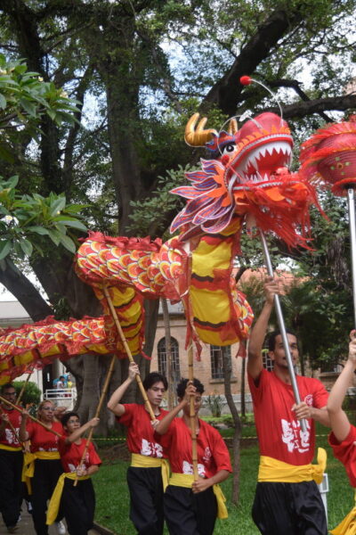 Nove pessoas vestindo camisetas vermelhas e faixas amarelas executam dança do dragão em caminho de parque com árvores e gramado. Dragão vermelho e amarelo é sustentado por varas, com uma participante segurando uma esfera vermelha na frente.